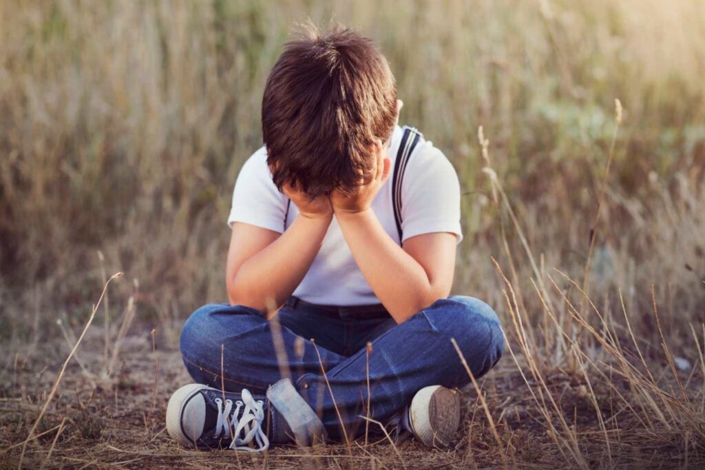 Junge sitzt geknickt in einer Wiese am Boden, sein Gesicht ist in seinen Händen vergraben. – Wie gelingt der Familienausflug? 5 Tipps!
