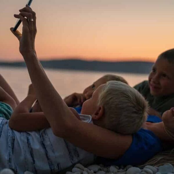 Frau liegt am Boden, mit ihren 3 Kindern im Arm und alle schauen auf ein Smartphone - Tipps für Ferien-Apps