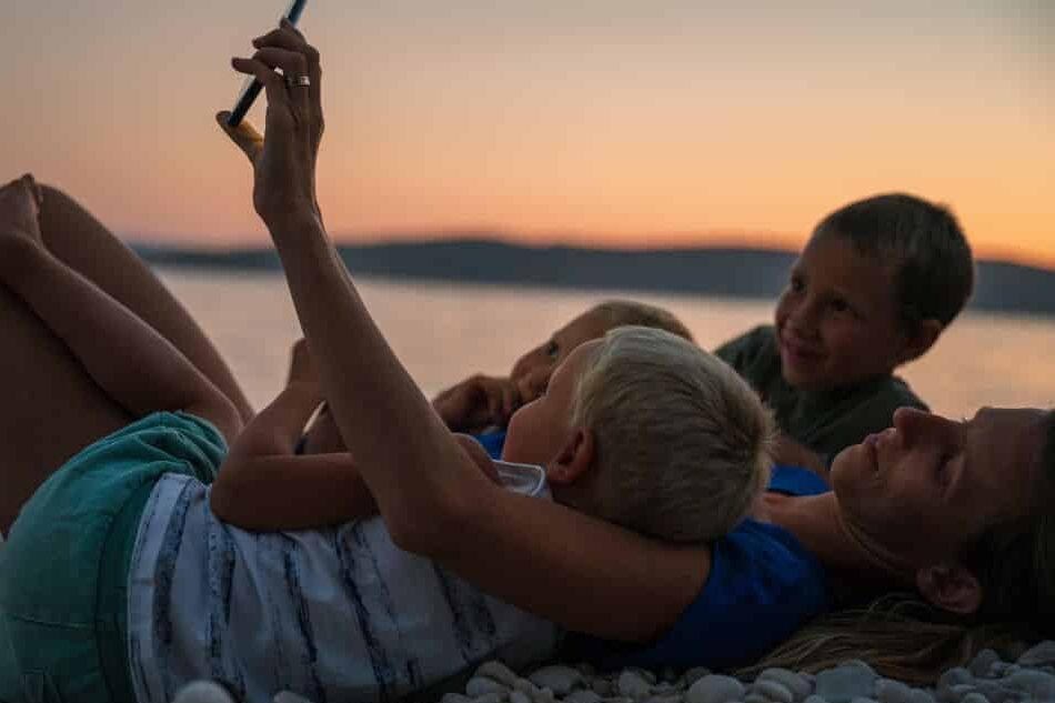 Frau liegt am Boden, mit ihren 3 Kindern im Arm und alle schauen auf ein Smartphone - Tipps für Ferien-Apps