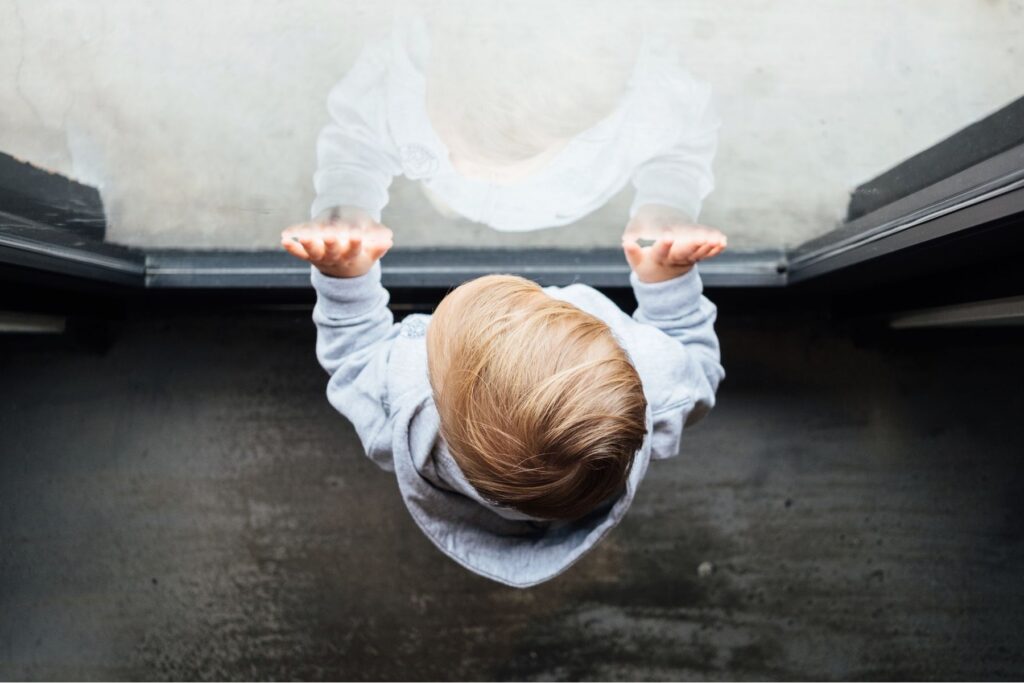 Kind, von oben fotografiert, lehnt an einer Glasscheibe und schaut hinaus - Trennungsangst: Was tun, wenn das Kind beim Abschied weint?