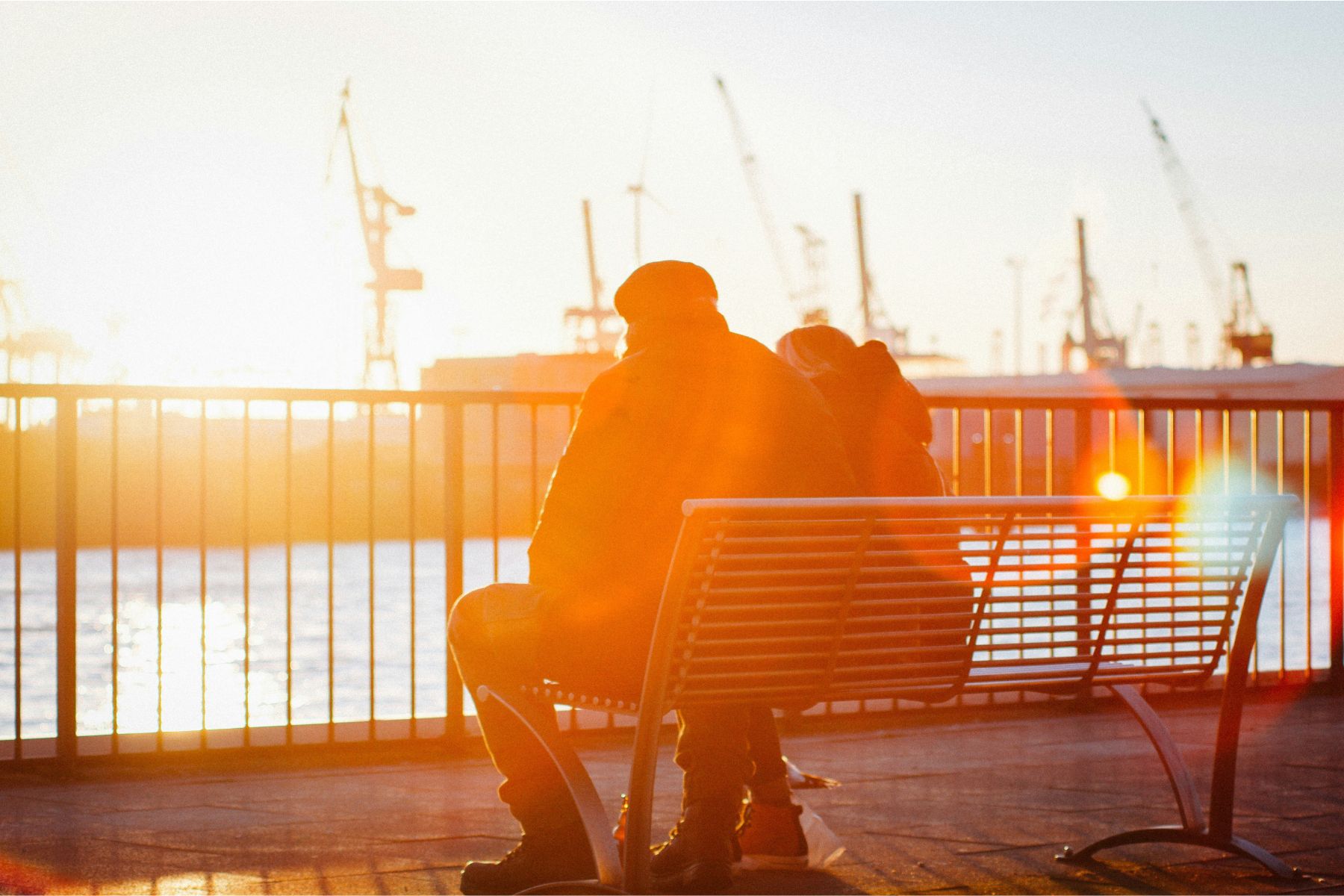 Ein älteres Paar mit Rücken zur Kamera auf einer Bank am Wasser sitzend, bei Sonnenuntergang.