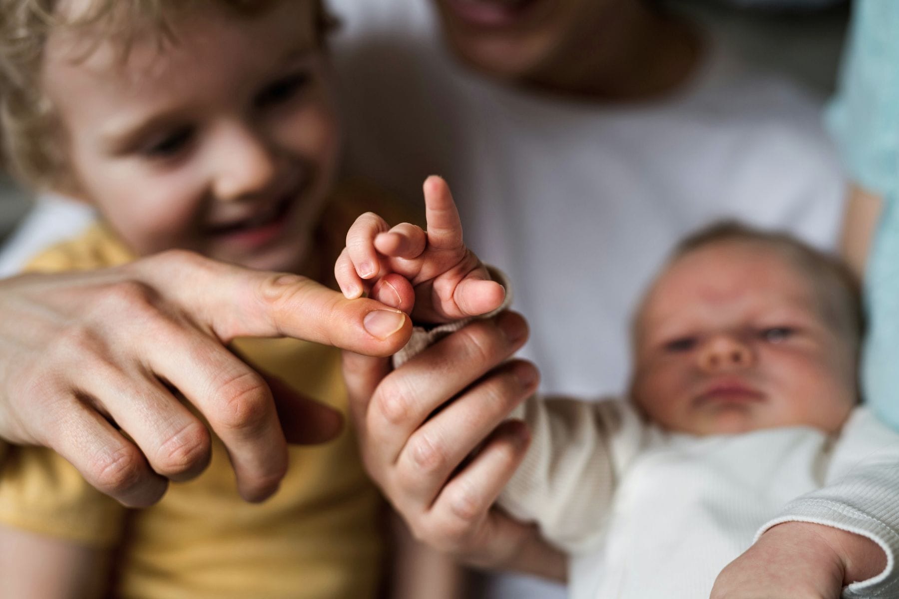 Eine Babyhand, gehalten von den Händen eines Erwachsenen, daneben ist unscharf ein älteres Geschwisterkind erkennbar. – Das zweite Kind.