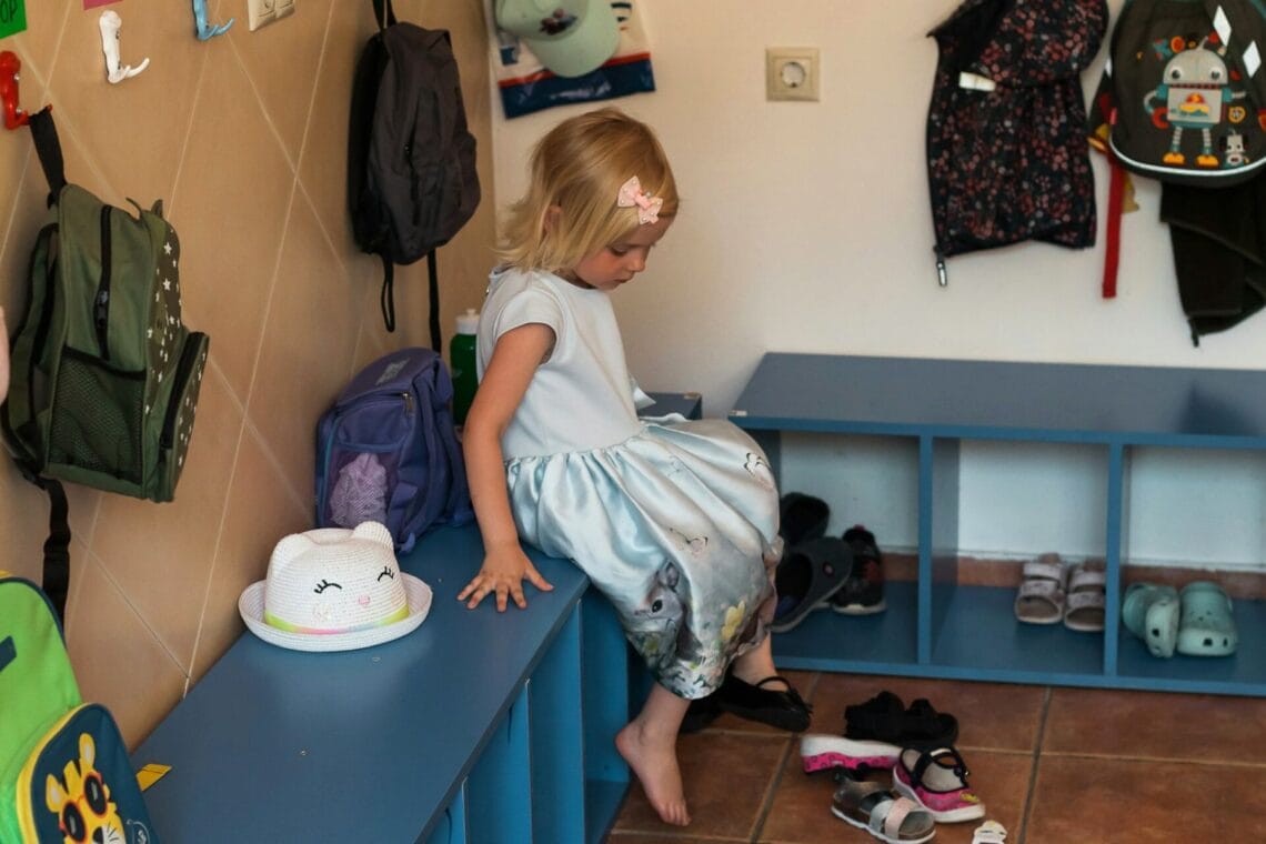 Kleines Mädchen sitzt in der Kindergarten-Garderobe und wechselt von Schuhen auf Finken. Unsere Empfehlungen für den Kindergartenstart.