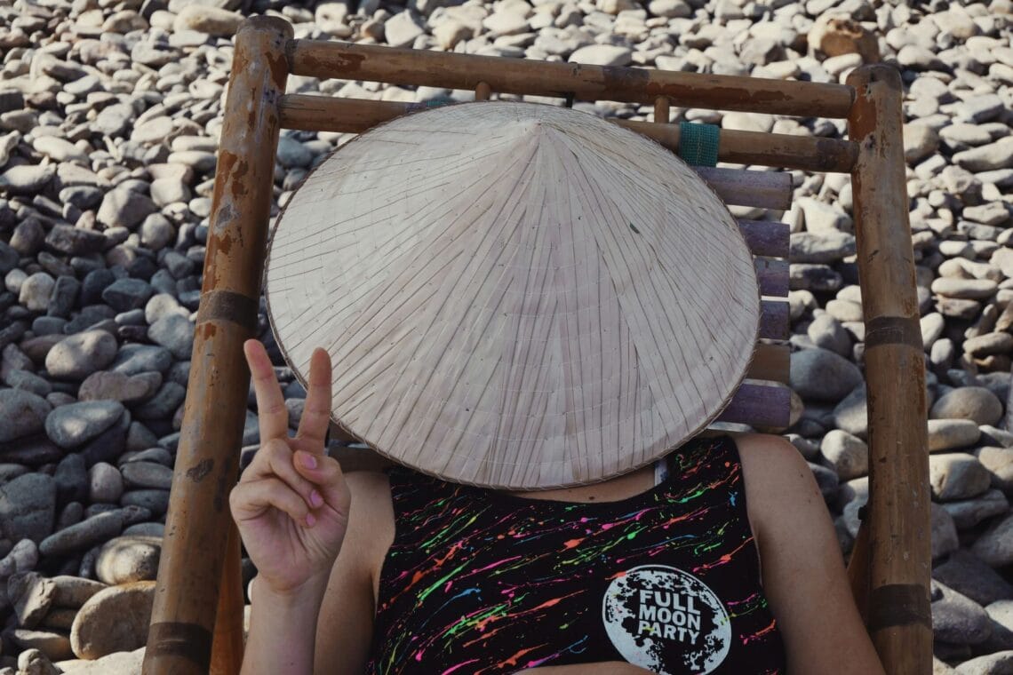 Teenagerin liegt auf hölzernem Liegestuhl am Kiesstrand, ihr Gesicht ist mit einem Strohhut bedeckt, ihre Finger formen das Peace-Zeichen.
