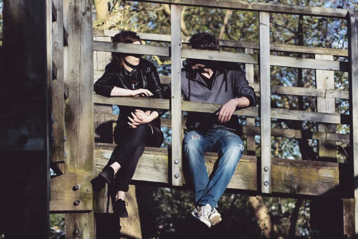 Frau und Mann sitzen Hände haltend auf einer Holzbrücke und schauen sich an. Hilft ehrliche Kommunikation gegen emotionalen Beziehungsfrust?