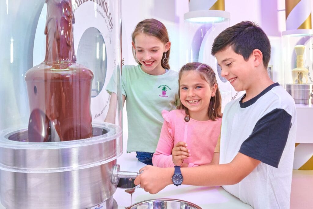 Drei Kinder stehen lächelnd neben einem Schokoladenbrunnen im Chocolarium Flawil - Ostschweiz mit Kindern bei Regen - unsere Tipps.