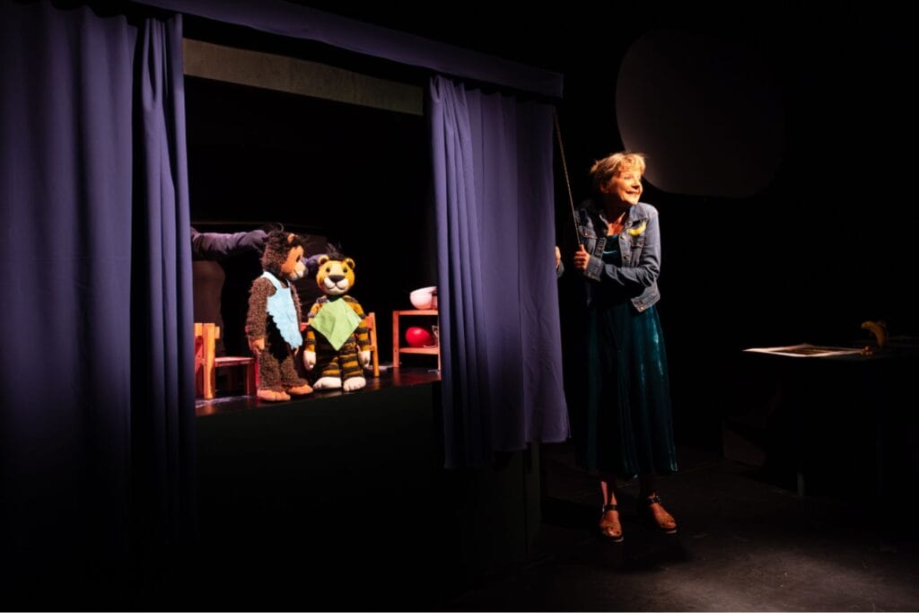 Eine Frau öffnet einen blauen Vorhang. Auf der Bühne sind zwei Tierfiguren zu sehen. – Erprobte Tipps für Ostschweiz mit Kindern bei Regen