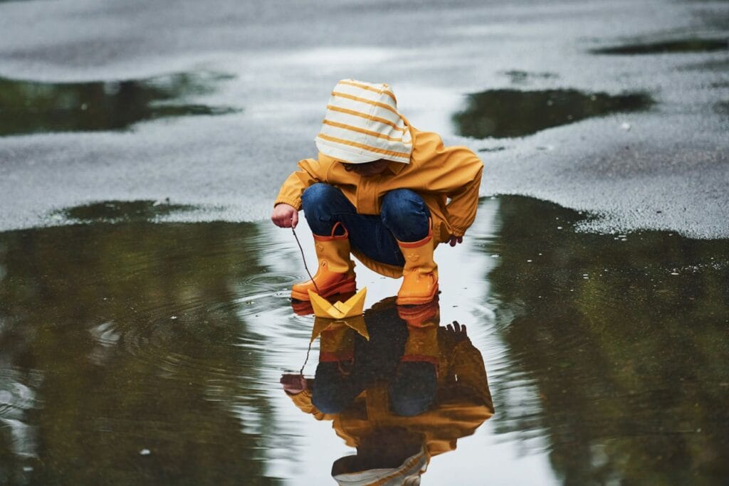 Kleinkind kauert in einer Pfütze und schiebt mit einem Ast ein Papierschiff an. Was tun in der Ostschweiz mit Kindern bei Regen?Unsere Tipps