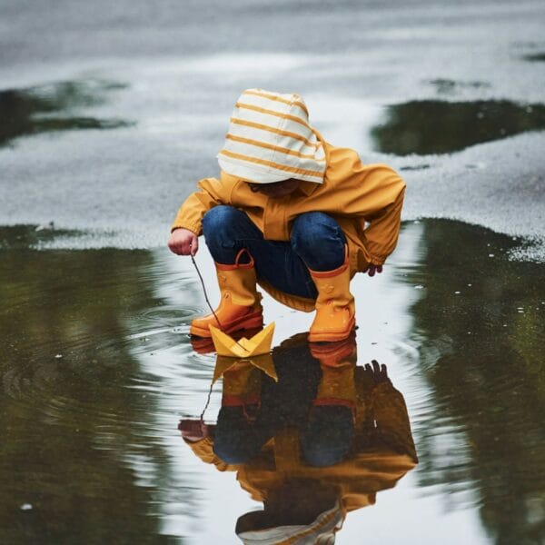 Kleinkind kauert in einer Pfütze und schiebt mit einem Ast ein Papierschiff an. Was tun in der Ostschweiz mit Kindern bei Regen?Unsere Tipps