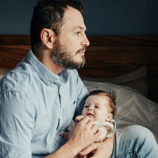 Mann sitzt auf Bett, hält Baby im Arm und schaut nachdenklich in die Ferne. – Alex will ein engagierter Vater sein - und landet im Burn-out.