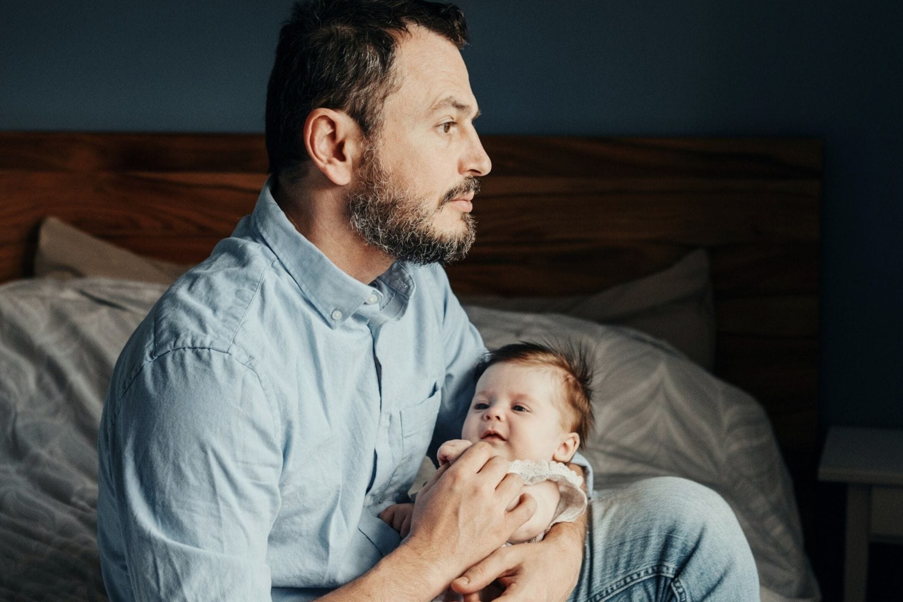 Mann sitzt auf Bett, hält Baby im Arm und schaut nachdenklich in die Ferne. – Alex will ein engagierter Vater sein - und landet im Burn-out.