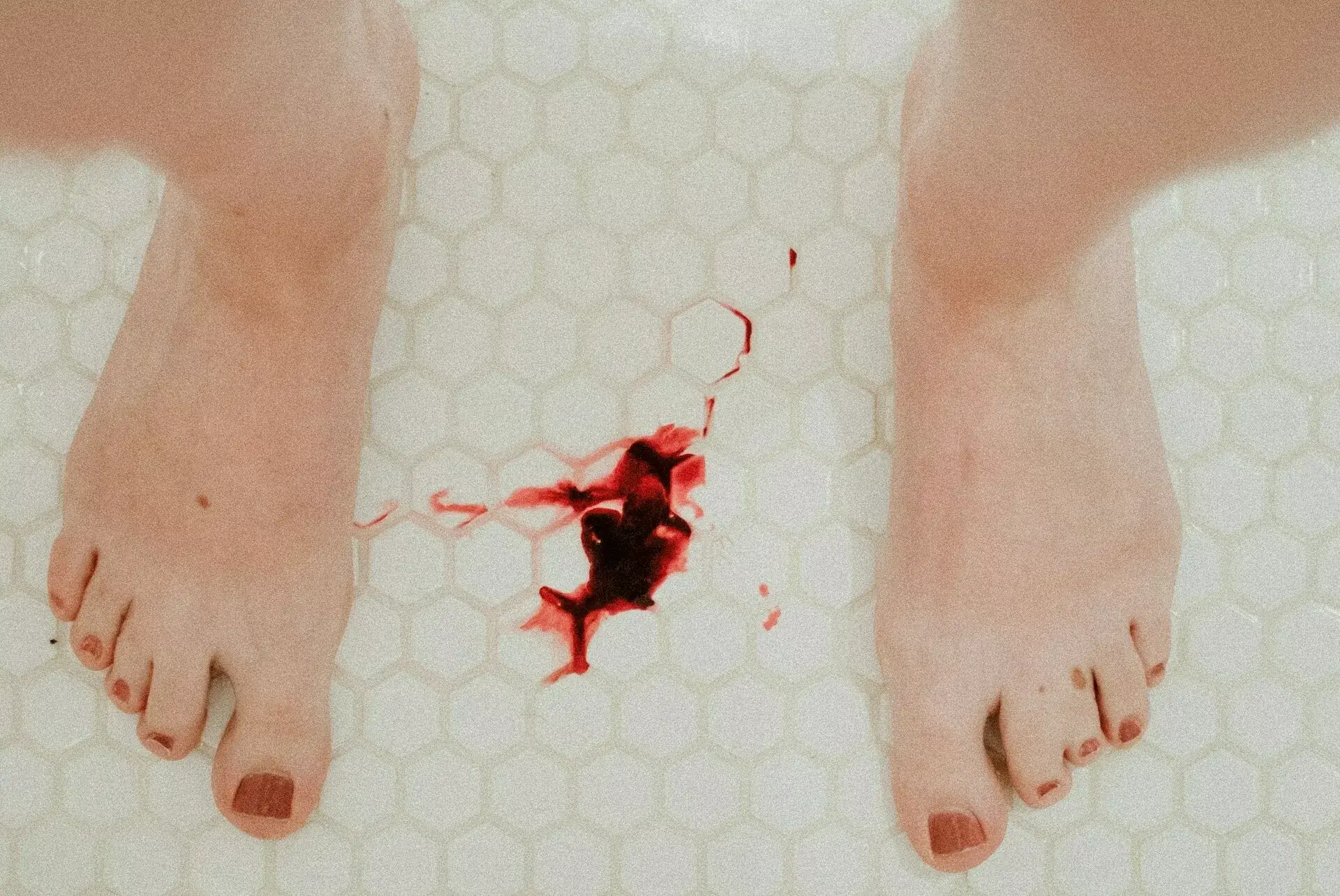 Blutfleck auf Badezimmerplättli, umrahmt von nackten Frauenfüssen mit rot lackierten Nägeln. - Wie wir unseren Zyklus für uns nutzen können.