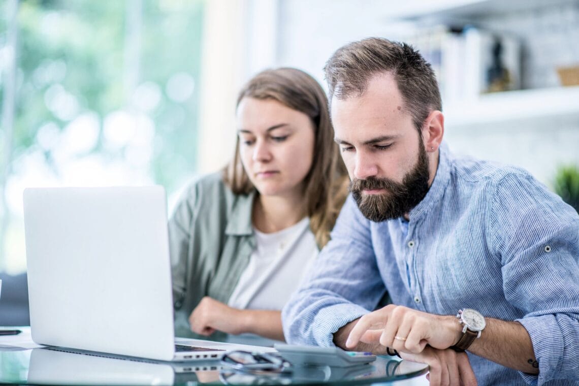 Frau und Mann sitzen am Tisch und schauen auf Laptop und Taschenrechner. Über Geld wird in Paarbeziehungen wenig geredet und oft gestritten.