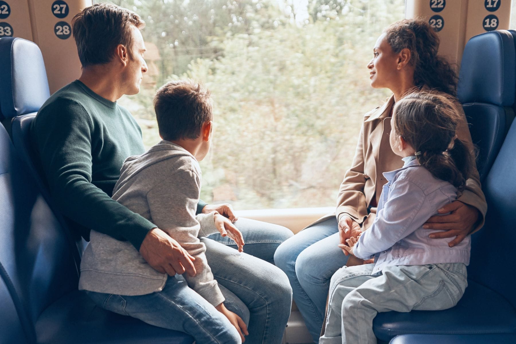Familie mit Mutter, Vater, Tochter, Sohn sitzt im Zugabteil und schaut aus dem Fenster. Familienferien mit dem Zug? Eine Expertin gibt Tipps