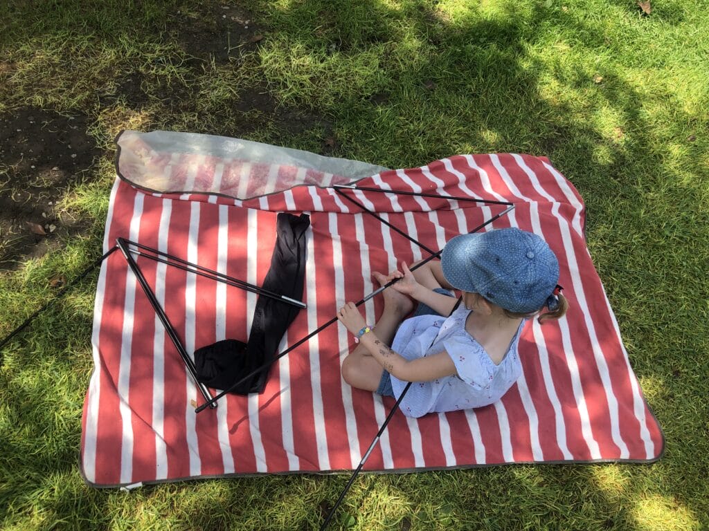 Kleines Mädchen sitzt auf einer Picknickdecke und hantiert mit den Zeltstangen. Die besten Campingplätze für Familien – unsere Empfehlungen