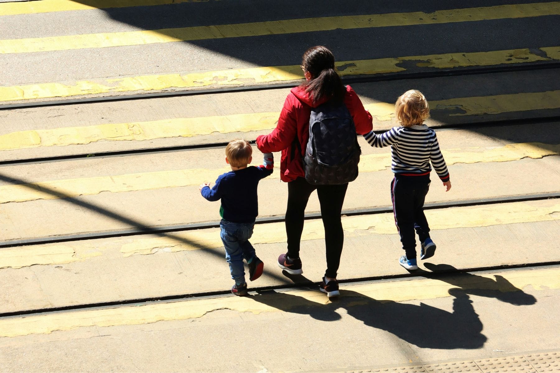 Eine Frau läuft mit zwei Kindern an der Hand über einen Fussgängerstreifen. Die Personen werfen lange Schatten. - alleinerziehend