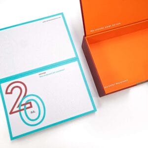 Aufgeklapptes Kalenderblatt mit Schreibimpuls neben der offenen Box mit orangem Innenleben
